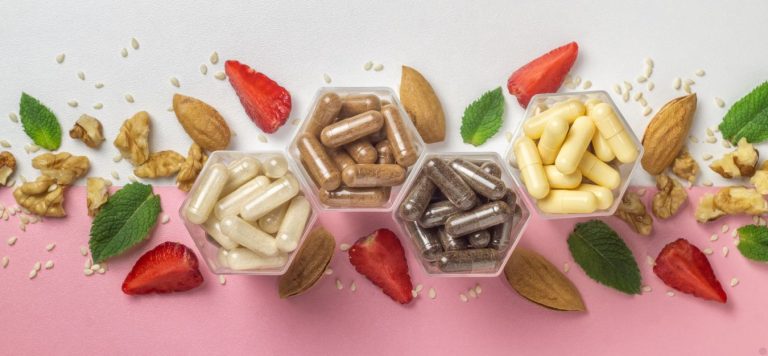 Different vitamins on a white and pink background, with strawberries and nuts laid out around them.