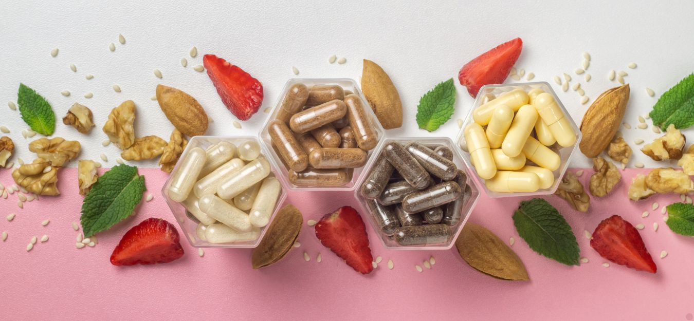 Different vitamins on a white and pink background, with strawberries and nuts laid out around them.